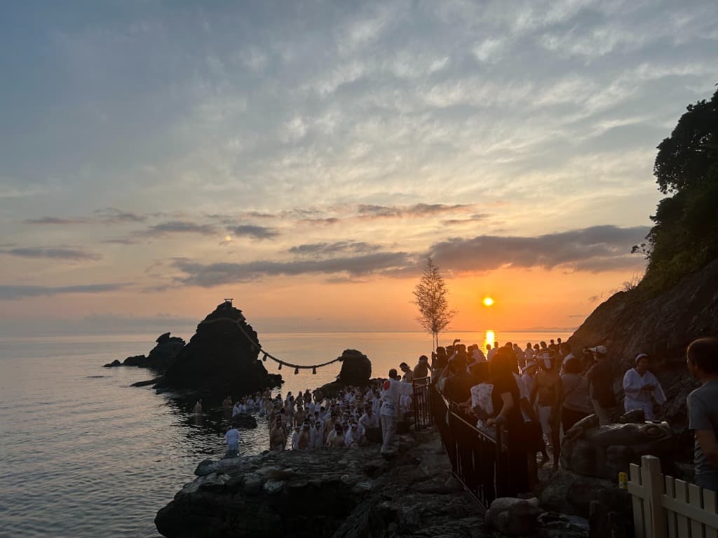 夫婦岩と水平線に沈む夕日。参拝者が岩場に並んでいる